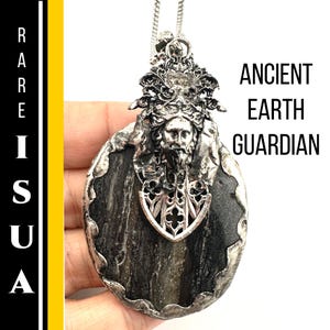 Puede incluir: Un collar con colgante de plata con un diseño de Guardián de la Tierra Antigua. El colgante tiene una piedra oscura de forma ovalada con una figura plateada detallada y adornos. El texto "ANCIENT EARTH GUARDIAN" es visible.
