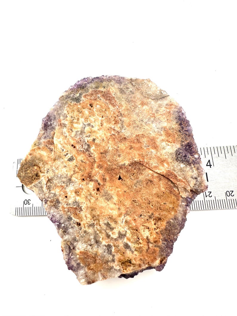 May include: A close-up of a purple and brown agate stone with a rough, natural texture. The stone is approximately 4 inches wide and is sitting on a white surface.