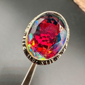 May include: An oval-shaped ring featuring a large, faceted, iridescent red gemstone with multi-coloured light reflections. The stone is set in a silver-toned metal band with a decorative, textured, and engraved border surrounding the setting.