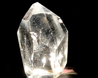 May include: A clear quartz crystal with a faceted, pointed shape. The crystal is transparent, allowing light to pass through, and sits against a black background. The crystal's surface has a slightly frosted appearance.