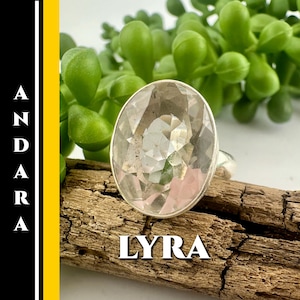 Pode incluir: Um anel de prata com uma grande gema oval facetada. A gema é transparente com tons de rosa e está sobre um pedaço de madeira. A palavra "LYRA" é exibida em texto branco. A palavra "ANDARA" é exibida verticalmente em texto preto.