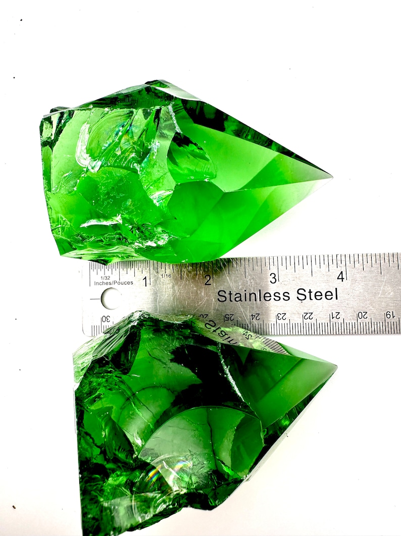 May include: Two large, green, faceted crystal points, one partially resting on a stainless steel ruler marked in inches. The ruler reads "Stainless Steel" and has markings from 1 to 4 inches.