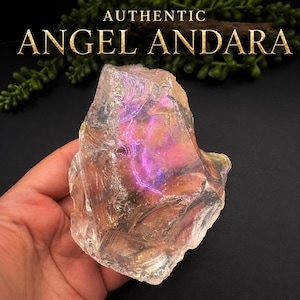 May include: A translucent, irregular-shaped crystal with a shimmering, iridescent surface displaying flashes of pink, purple, and gold. The text AUTHENTIC ANGEL ANDARA is printed in gold lettering above the crystal against a dark background.