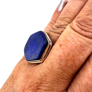 Puede incluir: Un anillo de plata con una gran piedra hexagonal de lapislázuli azul oscuro. El anillo se lleva en un dedo.