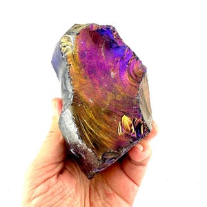 Puede incluir: Una gran piedra de obsidiana iridiscente con una superficie pulida. La piedra muestra una vibrante gama de colores, incluyendo púrpura, dorado y azul, con un brillo metálico reflectante. Los bordes ásperos contrastan con el interior liso y colorido.