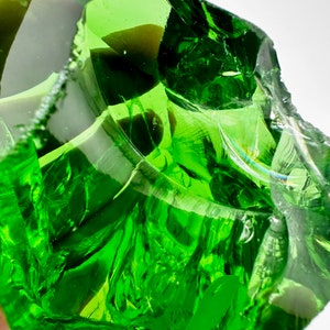 May include: A large, green, irregularly shaped piece of glass with a smooth, polished surface. The glass is translucent and has a slight green tint. The glass is reflecting light, creating a shimmering effect.