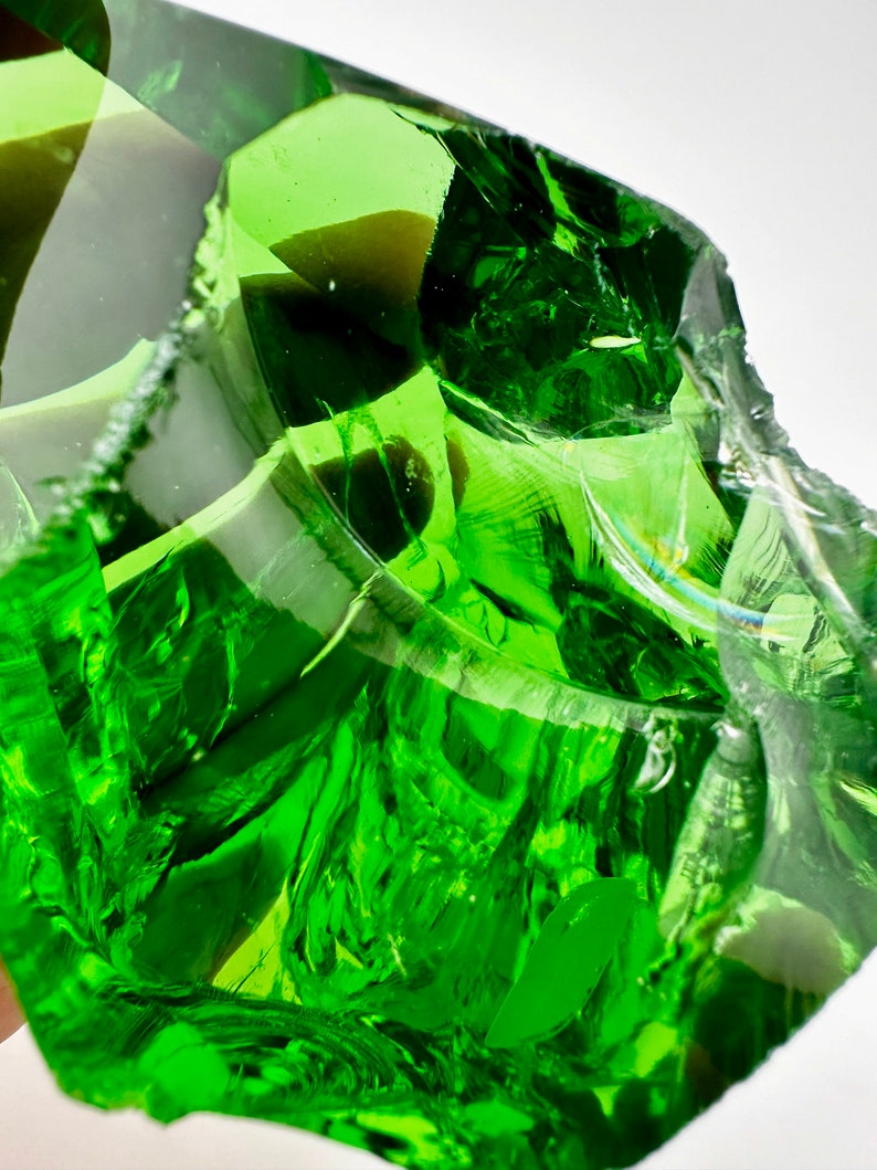 May include: A large, green, irregularly shaped piece of glass with a smooth, polished surface. The glass is translucent and has a slight green tint. The glass is reflecting light, creating a shimmering effect.