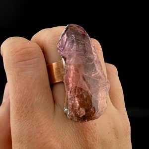May include: A large, raw amethyst crystal ring. The ring features a copper band and a large, irregular-shaped purple crystal. The crystal has a translucent quality, with variations in color from light to dark purple. The ring is set against a black background.