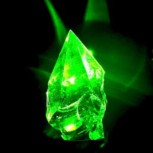May include: A bright green, faceted crystal with a pointed tip, illuminated from within, against a black background.
