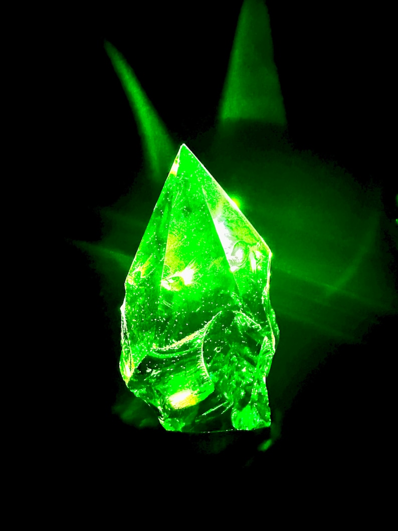 May include: A bright green, faceted crystal with a pointed tip, illuminated from within, against a black background.