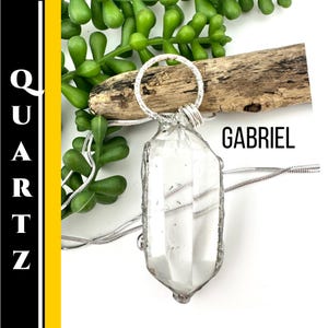 Puede incluir: Un collar con colgante de cristal de cuarzo transparente con un lazo y cadena de metal plateado. La palabra "GABRIEL" se muestra en texto negro. La palabra "QUARTZ" se muestra verticalmente en texto blanco sobre un fondo negro y amarillo.