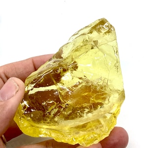 May include: A large, irregularly shaped, translucent yellow crystal. The crystal has a rough, fractured surface and is held in a hand against a white background. The color is a vibrant, sunny yellow.