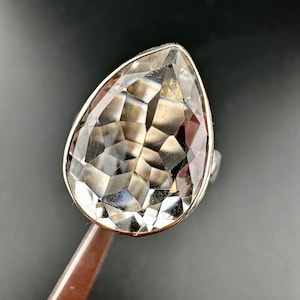May include: A silver ring featuring a large, pear-shaped, faceted gemstone. The clear stone has multiple internal facets, creating a sparkling effect. The ring's band is a simple, polished silver.