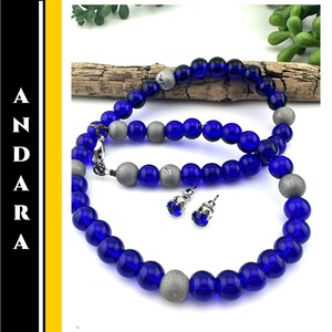 May include: A necklace and matching earrings set featuring deep blue glass beads and silver-toned accents. The necklace is composed of round, translucent beads interspersed with larger, matte silver beads. The earrings have blue gemstone studs. The word "ANDARA" is printed vertically on the left side.