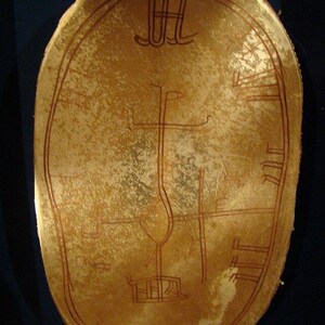 May include: A tan, oval-shaped artifact with intricate, reddish-brown line drawings. The design includes a central vertical line, a curved shape, and several horizontal and vertical lines, possibly depicting a map or symbolic representation. The object has a textured surface.