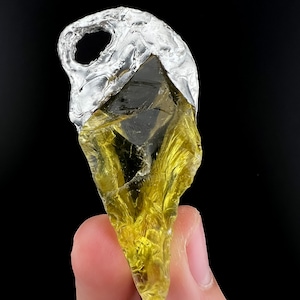 Puede incluir: Un colgante de piedra preciosa en bruto, de color amarillo verdoso, con una tapa y un lazo de metal plateado. El colgante tiene una textura natural rugosa, sobre un fondo negro.