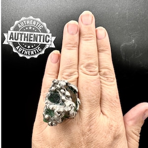 Puede incluir: Un anillo de plata con una piedra grande, verde y plateada. El anillo se lleva en el dedo de una persona.