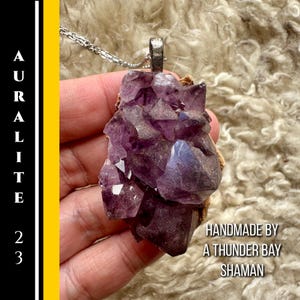 May include: Auralite 23 pendant necklace with a large, raw amethyst crystal in shades of purple and violet. The crystal is mounted on a silver-tone bail and hangs from a delicate chain. The text "HANDMADE BY A THUNDER BAY SHAMAN" is visible.
