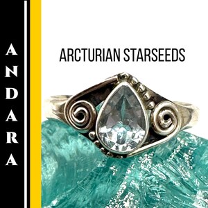 May include: A silver ring with a pear-shaped, light blue gemstone. The ring has a decorative swirl design on the sides and small beaded accents. The text "ARCTURIAN STARSEEDS" is displayed above the ring.