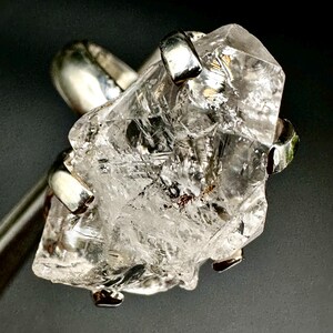 Puede incluir: Un anillo de cristal de cuarzo transparente con una banda de plata. El cristal está facetado y tiene un aspecto áspero y natural. El anillo es una pieza de joyería única y llamativa.
