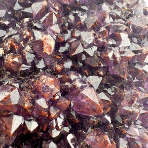 May include: A close-up of a cluster of amethyst crystals. The crystals are a deep purple color and have a natural, rough texture. Some of the crystals have a reddish-brown hue.
