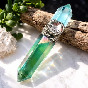 May include: A double-terminated crystal wand featuring a translucent green and blue iridescent finish. The centre is wrapped in a textured, antique-style silver metal band that holds a small, clear, round faceted gemstone in the middle.