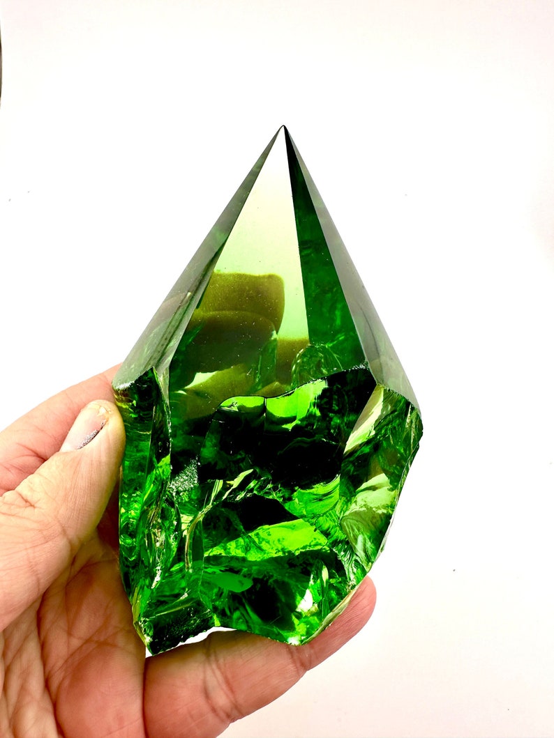 May include: A large, green, faceted glass crystal in the shape of a pyramid. The crystal is clear and has a slight yellow tint. It is being held in a hand.