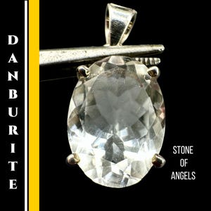 May include: A clear, faceted danburite pendant with a silver bail. The gemstone is held by four silver prongs. The text "DANBURITE" is vertically aligned on the left, and "STONE OF ANGELS" is on the right.