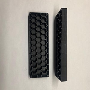 Peut inclure: Deux entretoises en plastique noir à motif nid d'abeille. Les entretoises sont rectangulaires et ont un bord surélevé autour du périmètre.