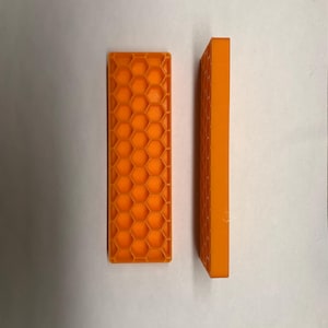 May include: Two orange rectangular objects. One is lying flat, displaying a honeycomb pattern. The other stands upright, showing a side view with a smooth surface. Both objects are made of a solid, opaque material.