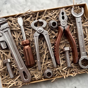 May include: A selection of silver and copper-coloured tools, including a spanner, screwdriver, pliers, and bolts, arranged in a brown box with shredded paper. The spanner has the words "CHROME VANADIUM" printed on it.