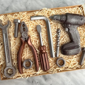 May include: A selection of tools, including a spanner, pliers, screwdriver, and drill, presented in a cardboard box with wood shavings. The tools are metallic grey and brown, creating a realistic appearance. The box is set against a light background.
