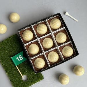 May include: A box of nine golf ball-shaped chocolates on a brown base, arranged in a grid. A white tee and a green flag with the number 18 are also present. Several golf ball chocolates are scattered around the box.