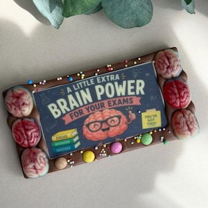 May include: A chocolate bar decorated with brain-shaped sweets and sprinkles. The centre has a blue graphic with the words "BRAIN POWER FOR YOUR EXAMS". The treat is designed to look like a brain.