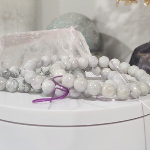 May include: Two white stone beaded bracelets with purple string ties. The bracelets are on a white circular display stand.