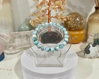 May include: A light blue gemstone bracelet with a round shape. The bracelet is made of many small beads and is displayed on a white stand.
