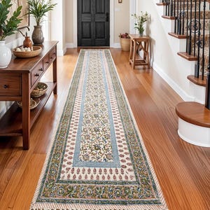 Könnte beinhalten: Ein langer, rechteckiger Teppich mit cremefarbenem Hintergrund und einem Blumenmuster in Blau-, Grün- und Rottönen. Der Teppich hat einen Rand mit sich wiederholendem Design und Fransen an den Enden. Er liegt auf einem Holzboden.