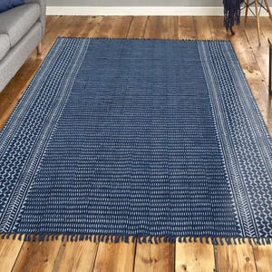Puede incluir: Una alfombra rectangular azul marino con un borde geométrico blanco y flecos. La alfombra presenta un patrón repetitivo de pequeños guiones blancos y formas geométricas. La alfombra está colocada sobre un suelo de madera en un salón.