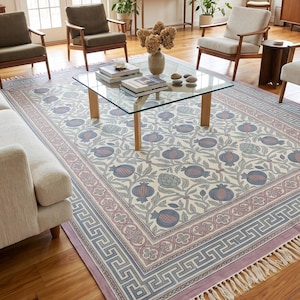 Puede incluir: Una alfombra rectangular con fondo color crema y un patrón repetitivo de granadas azules y rosas con follaje verde. El borde presenta un diseño geométrico de greca griega en azul y rosa, rematado con flecos blancos en los extremos.