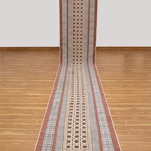 Puede incluir: Una alfombra larga y rectangular con un patrón geométrico. La alfombra presenta un diseño central de formas de diamante repetidas y un borde de rayas y patrones alternos. La paleta de colores incluye tonos de marrón, beige y blanco.