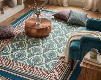 Op de afbeelding: Een rechthoekig vloerkleed met een turquoise en crèmekleurig bloemmotief, omzoomd door een geometrisch ontwerp en een turquoise franje. Een koperkleurige salontafel staat op het kleed, met een turquoise fluwelen stoel en kussens.
