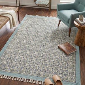 Puede incluir: Alfombra rectangular con estampado floral en tonos azules y crema. Tiene un borde azul claro y flecos. Un libro y zapatillas están sobre la alfombra. Ideal para el salón.