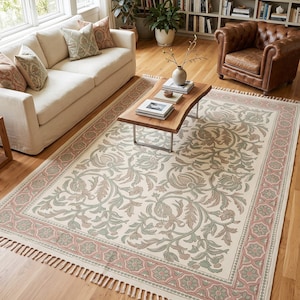 Puede incluir: Una alfombra rectangular con base de color crema y un patrón repetitivo de flores y enredaderas en tonos verde salvia y rosa empolvado. La alfombra tiene un borde decorativo ancho con motivos florales y flecos en ambos extremos.