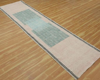 May include: A long, rectangular runner rug with a pink background and a geometric pattern of two overlapping squares in shades of green and blue. The rug has a black and white border with a decorative, jagged design.