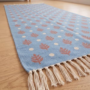 Puede incluir: Una alfombra rectangular de pasillo en color azul claro con un patrón repetitivo de pequeñas hojas de helecho color terracota y puntos crema. La alfombra tiene una textura tejida y está rematada con flecos color crema en los bordes cortos.