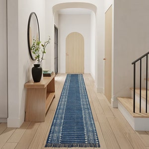 May include: A long, navy blue and white patterned runner rug with a fringed edge. The rug features a geometric design with rows of triangles and dots. It is placed in a hallway with light wood flooring.