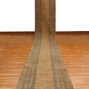 May include: A long, narrow, brown rug with a black and white geometric border. The rug is laid out on a wooden floor.
