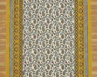 Op de afbeelding: Een rechthoekig vloerkleed met een crèmekleurig middenstuk met een paisley patroon in bruin- en blauwtinten. Het vloerkleed heeft een gele rand met een bloemenmotief en franjes aan de korte uiteinden.