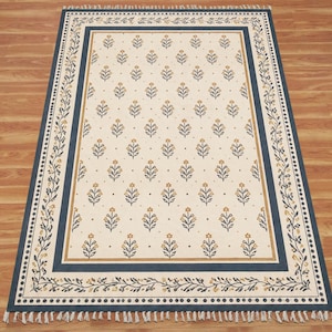 Pode incluir: Um tapete retangular com um centro de cor creme apresentando um padrão repetitivo de pequenas flores amarelas e caules azul-escuros. O tapete é emoldurado por uma borda azul-escura com um motivo de videira floral e franjas brancas nas pontas.