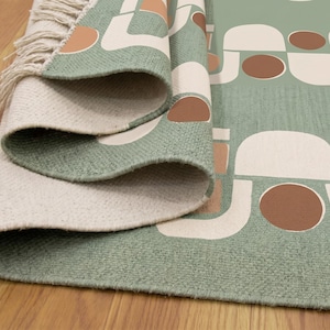 Puede incluir: Una alfombra enrollada con un patrón geométrico en verde salvia, crema y marrón. La alfombra tiene un borde con flecos y se exhibe sobre un piso de madera. El diseño presenta formas abstractas.